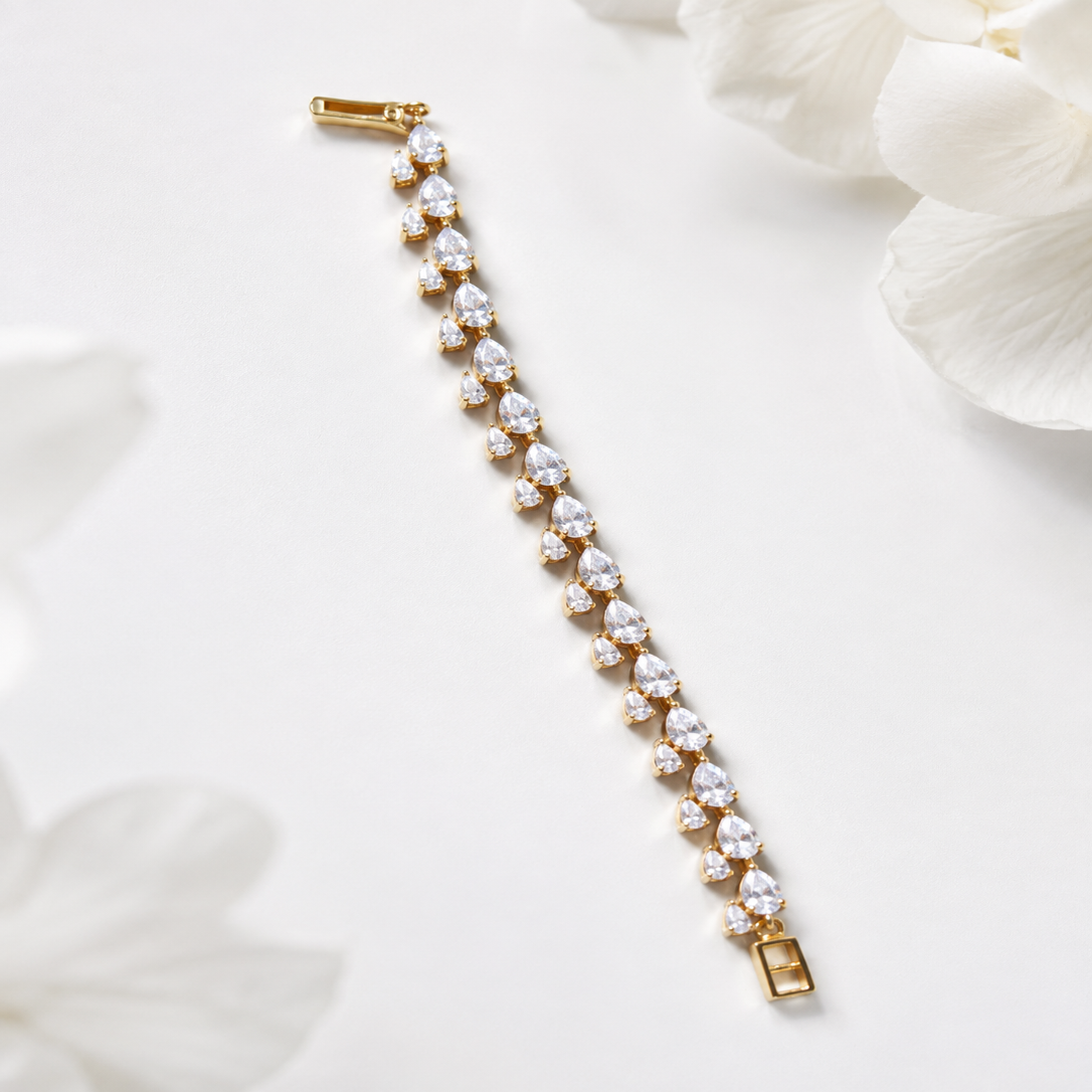 A gold tennis bracelet with clear teardrop-cut gemstones, laid diagonally on a white background with soft white flower petals in the corners.