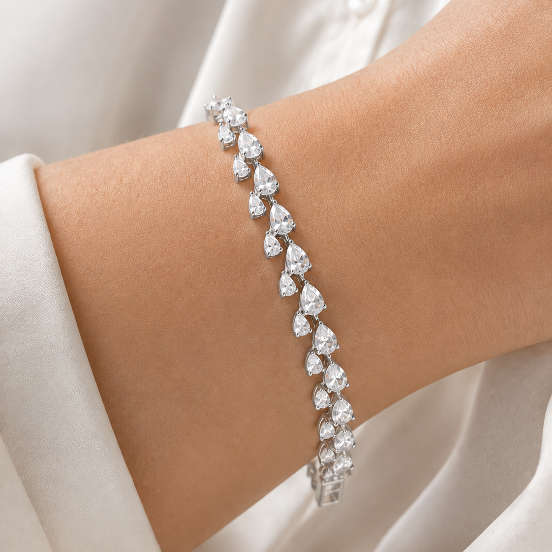 A silver tennis bracelet with heart-shaped clear gemstones worn on a woman's wrist, set against a soft white blouse.